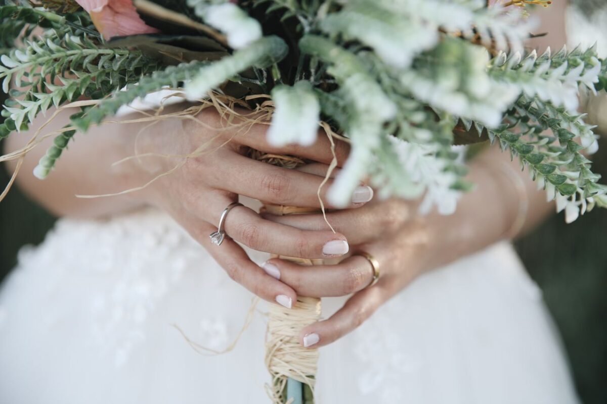 A bride's hands holding a beautiful floral wedding bouquet, highlighting rings and intricate florals.