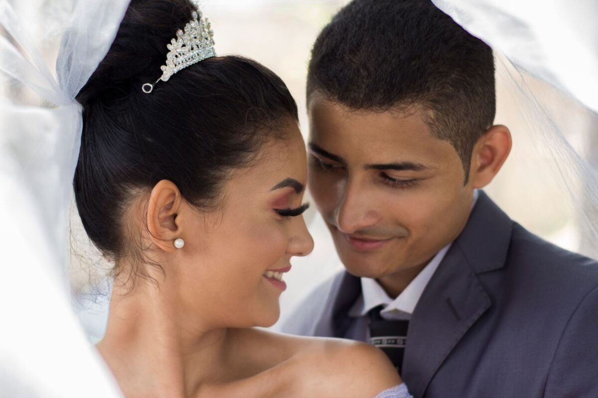 Ethnic groom standing near bride with white veil and diadem on head while looking at each other during wedding ceremony