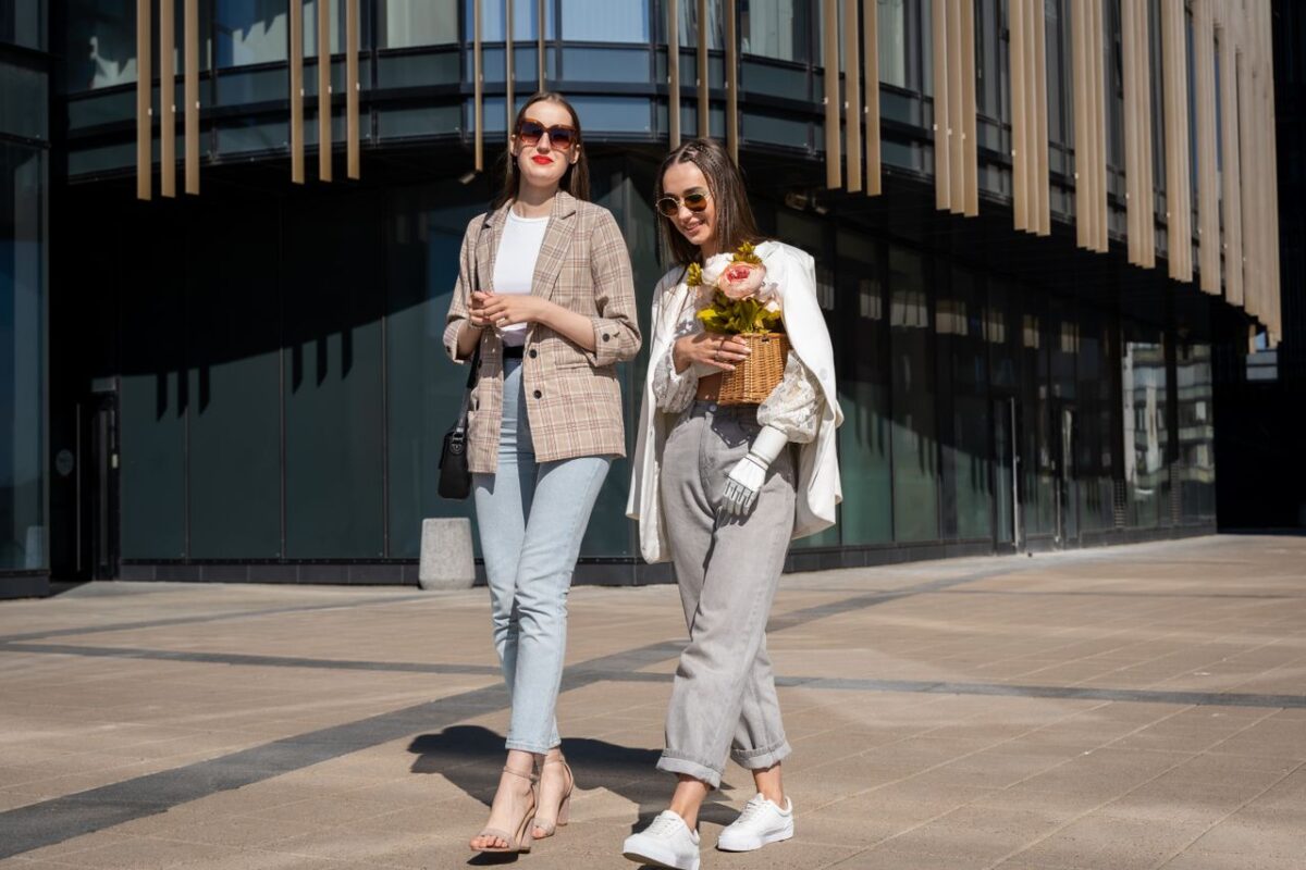 Two fashionable women with sunglasses, one holding flowers, casually walking outdoors, showcasing urban lifestyle.